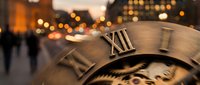 Close-up of vintage clock face showing Roman numerals with blurred city street lights and pedestrians in warm evening atmosphere for time concepts.