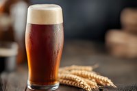 Cold glass of dark craft beer with foam head and water drops alongside golden wheat ears on rustic wooden table, moody dark background.