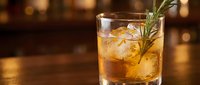 Golden whiskey cocktail with ice cubes and fresh rosemary garnish on wooden bar counter with warm bokeh lights creating elegant drinking atmosphere.