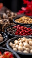 Assorted spices and dried ingredients in black ceramic bowls including red chili peppers, cloves and nuts on dark wooden surface with dramatic lighting.