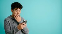 Young man with curly dark hair and olive complexion holding smartphone, wearing gray hoodie, touching face with thoughtful expression against turquoise background for tech and communication concepts.