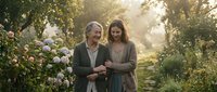 Mature man and woman strolling together through blooming garden pathway surrounded by white flowers and lush greenery during golden hour, conveying romance and companionship.