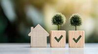 Wooden house model and blocks with green checkmarks and decorative trees, representing eco-friendly real estate and sustainable home concepts on blurred background.