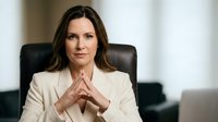 Professional businesswoman in beige suit sitting at office desk with hands clasped, confident executive portrait for corporate communications and leadership content.