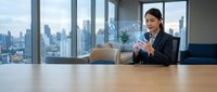 Professional Asian businesswoman using smartphone in modern office conference room with city skyline view through large windows.