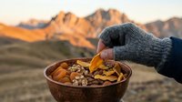 Hand holding bowl of mixed nuts and dried fruit against mountain landscape at sunset, perfect for outdoor adventure, healthy snacking, and travel lifestyle content.