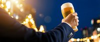 Man in dark business suit holding glass of beer in toast against blurred city lights at night, celebrating success or business achievement.