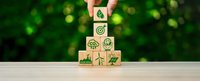 Hand placing wooden cube with green eco symbols forming pyramid on white surface against blurred natural background for environmental awareness campaigns.