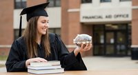 Young Caucasian female graduate in cap and gown holding piggy bank with stack of books, symbolizing education investment and financial planning.