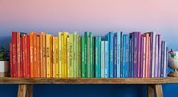 Rainbow collection of colorful books arranged by color spectrum on wooden shelf against pink wall creating organized library display for education and reading.
