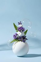 White ceramic vase with purple flowers and green fern leaves surrounded by floating soap bubbles on soft blue background for spa wellness decor.