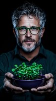 Mature man with gray hair and green glasses holding bowl of fresh microgreens sprouts on dark background. Professional portrait for organic food business.