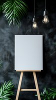 Blank white canvas on wooden easel against dark textured wall with vintage Edison bulbs and tropical palm leaves creating artistic atmosphere.