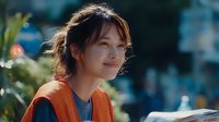 Young Asian woman with gentle smile wearing orange vest and gray shirt outdoors, reading document in bright natural light with blurred urban background.