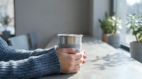 Woman hands holding steel mug with hot beverage in cozy home interior with plants and natural light creating peaceful morning atmosphere.