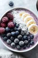 Purple smoothie bowl topped with fresh blueberries, raspberries, sliced banana and shredded coconut on marble background, healthy breakfast concept.