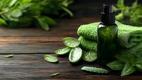 Natural aloe vera gel in dark glass bottle with fresh sliced leaves and green towel on wooden surface for skincare and wellness treatments.