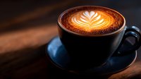 Hot cappuccino with latte art foam pattern in dark ceramic cup on wooden table. Moody coffee shop atmosphere with dramatic lighting and shallow depth of field.