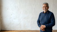Mature man with gray hair and glasses wearing dark blue sweater standing against white textured wall, smiling with hands clasped, suitable for corporate portraits and professional headshots.