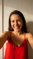 Happy young woman taking selfie in red dress with bright smile and natural sunlight creating warm shadows on white wall background for social media content.