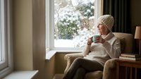 Young woman in cozy knit sweater and beanie enjoying hot beverage by window on snowy winter day, relaxing at home with warm drink and peaceful atmosphere.