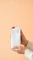 Hand holding white carton with red cap against peach background. Beverage packaging mockup for dairy, juice, or milk product advertising and branding purposes.
