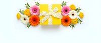 Yellow gift box with polka dot ribbon surrounded by colorful gerbera daisies and ranunculus flowers on white background, flat lay.