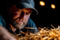 Dramatic portrait of bearded Caucasian farmer in cap examining fresh hay in barn with warm lighting creating moody atmospheric effect at dusk.