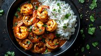 Spicy grilled shrimp in rich sauce served with steamed white rice and fresh herbs in black bowl on dark background. Homemade seafood dinner preparation.