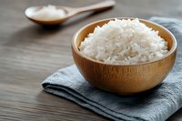 Steamed white jasmine rice served in wooden bowl on gray napkin, with wooden spoon aside. Natural light on rustic wooden table surface.