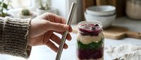 Person in beige sweater holding metal straw in glass jar with layered drink containing purple and green ingredients, sustainable lifestyle and eco-friendly beverage concept.