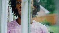 Young African American man with dreadlocks looking thoughtfully through window glass, reflecting on life decisions and future opportunities in modern home setting.