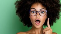 Young African American woman with natural afro hair and glasses showing surprised expression with raised finger against green background. Concept of idea or eureka moment.
