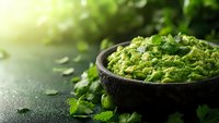 Fresh guacamole dip with cilantro in rustic stone bowl on dark background with natural sunlight and copy space. Mexican appetizer for healthy eating.