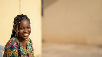 Young African woman with braided hair wearing colorful patterned clothing, smiling outdoors against neutral background. Suitable for diversity, lifestyle, and portrait projects.