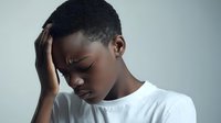 Young African American boy holding his head with hand, looking stressed and sad, wearing white t-shirt on light gray background.
