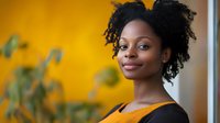 Young African American woman with natural curly hair smiling confidently against vibrant yellow wall, wearing black top. Professional portrait suitable for business or lifestyle content.