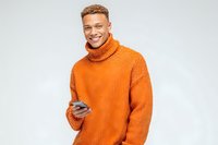 Happy young African American man in orange turtleneck sweater holding smartphone, smiling at camera on white background for lifestyle and technology concepts.