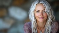 Confident mature woman with silver gray wavy hair and natural makeup smiling at camera, wearing purple knit sweater against blurred background.