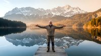 Middle-aged man with arms outstretched standing on wooden dock at mountain lake during golden hour sunrise with snow-capped peaks and autumn forest reflection.