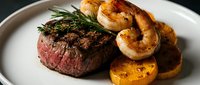 Grilled medium-rare steak with succulent shrimp, roasted potatoes, and fresh rosemary garnish on white plate for fine dining and restaurant menus.