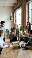 Diverse team of professionals gathered around wooden table in modern loft office with exposed brick, large windows, and plants, discussing business strategy and collaboration during casual meeting.