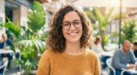 Happy young woman with curly hair and glasses smiling in modern office space with green plants and colleagues working in background during daytime.