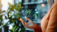 Caucasian woman using futuristic holographic interface with digital icons and data visualization, surrounded by green plants in modern office environment.