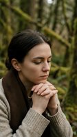 Peaceful young woman with closed eyes in prayer pose outdoors surrounded by lush green forest, expressing mindfulness and spiritual connection with nature.