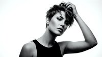 Androgynous young adult with short dark hair wearing black sleeveless top, hand raised to head in contemplative pose, studio portrait against white background for fashion and lifestyle content.
