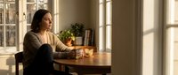 Young woman enjoying morning coffee at wooden table by sunny window in cozy home interior with natural light and plants creating peaceful atmosphere.