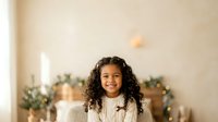 Young girl with long curly dark hair and warm smile wearing cream sweater, standing indoors with festive greenery and warm lighting in background.