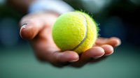 Hand holding bright yellow-green tennis ball against blurred court background, showcasing sports equipment for tennis match or practice.