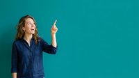 Young woman with long wavy brown hair, Caucasian ethnicity, wearing navy blue shirt, pointing upward with excited expression against turquoise background for advertising and promotional use.